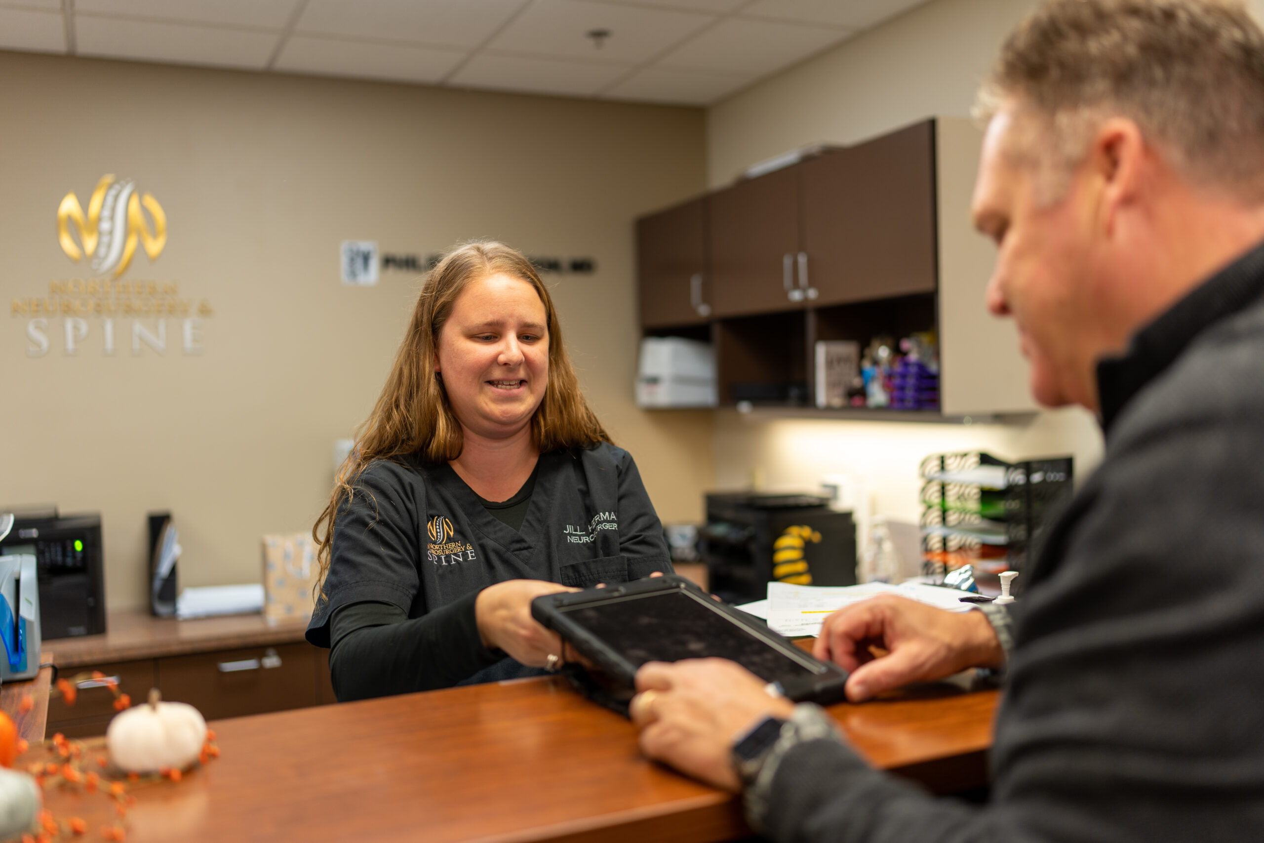A receptionist in a medical office hands a digital tablet to a man across the counter.