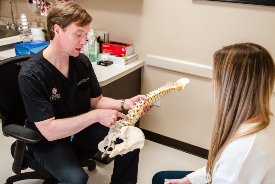 Man holding a full spine figure with pelvis, pointing to it, with woman in the foreground